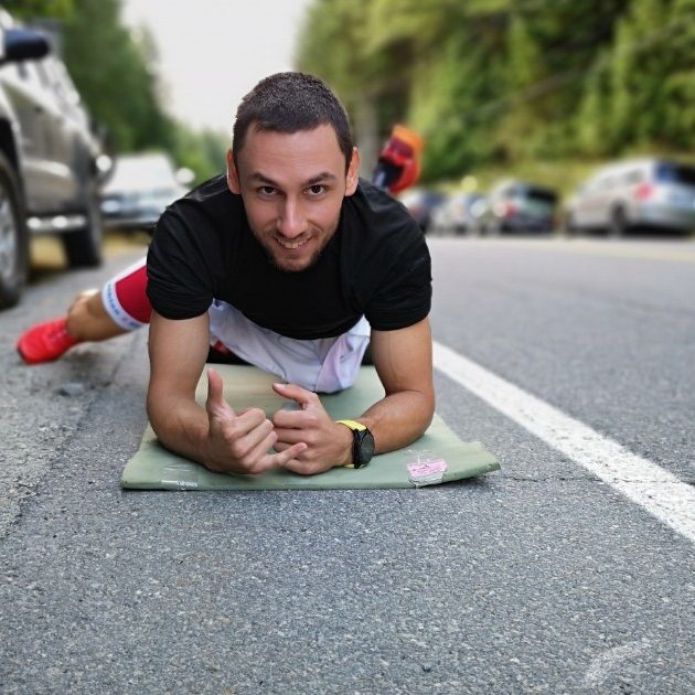 A man is in plank position on the ground facing the camera and smiling with his right leg out to the side.